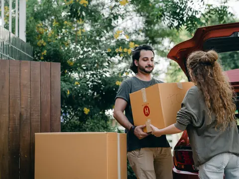 Two people passing a moving box to each other beside a car on a sunny street