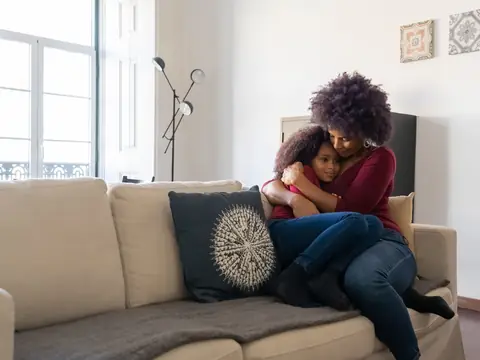 A mother holding her daughter close on a sofa in a sunny living room